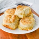 A white plate filled with golden, flaky biscuits sits on a wooden board—perfect for christmas brunch ideas. In the background, a dish of dark jam and a white napkin with a whisk can be seen.