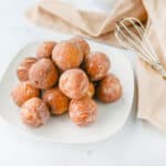 A white plate piled with homemade donut holes sits on a white surface next to a beige napkin and a metal whisk.