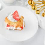A strawberry shortcake, one of the classic desserts with biscuits, is topped with whipped cream and a strawberry slice on a white plate, alongside measuring spoons, two spoons, a glass of milk, and a white cloth.