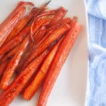 Brown sugar carrots roasted to perfection, cut into sticks and arranged on a white plate beside a light blue cloth with small white dots.