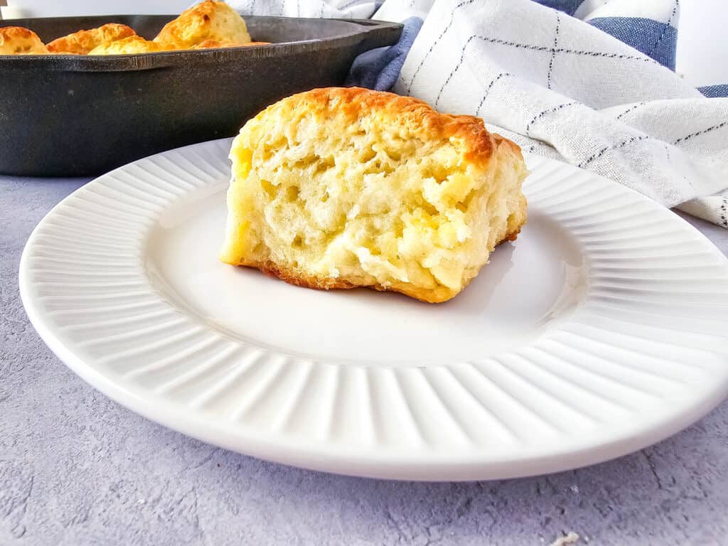 A golden, fluffy buttermilk pan biscuit sits on a white plate with a ridged edge. In the background, a cast iron pan with more biscuits and a white kitchen towel are visible.