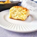 A golden, fluffy buttermilk pan biscuit sits on a white plate with a ridged edge. In the background, a cast iron pan with more biscuits and a white kitchen towel are visible.