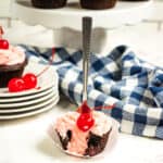 A chocolate cupcake with pink frosting and a cherry on top sits in a white wrapper. A fork is inserted into the cupcake, inspired by a Dr Pepper cupcakes recipe. Blue and white checkered cloth, plates, and more cupcakes are also visible.