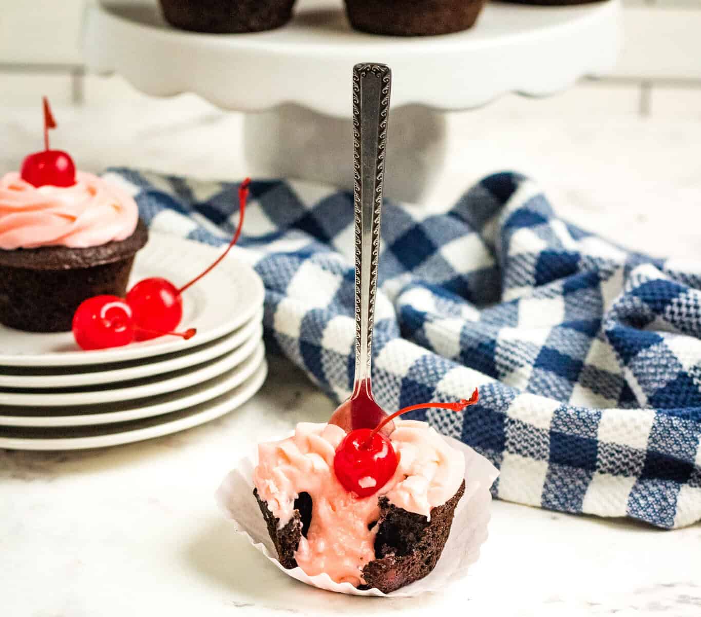 A chocolate cupcake with pink frosting and a cherry on top sits in a white wrapper. A fork is inserted into the cupcake, inspired by a Dr Pepper cupcakes recipe. Blue and white checkered cloth, plates, and more cupcakes are also visible.