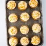 A metal baking tray holds twelve Butterbeer Cupcakes with swirled white frosting and yellow sprinkles, arranged in rows on a white surface.