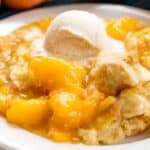 A serving of peach cobbler, a classic among desserts with biscuits, features a golden biscuit topping and a scoop of vanilla ice cream on a white plate. The juicy peaches glisten as the creamy ice cream begins to melt.