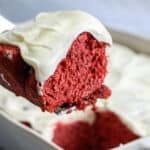 A close-up of a slice of red velvet cake with creamy white frosting being lifted from a baking dish filled with more frosted cake—perfect for a festive Valentines Breakfast for Kids.