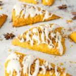 Several triangular pumpkin scones drizzled with white icing are arranged on a light surface, with a few star anise scattered around them.