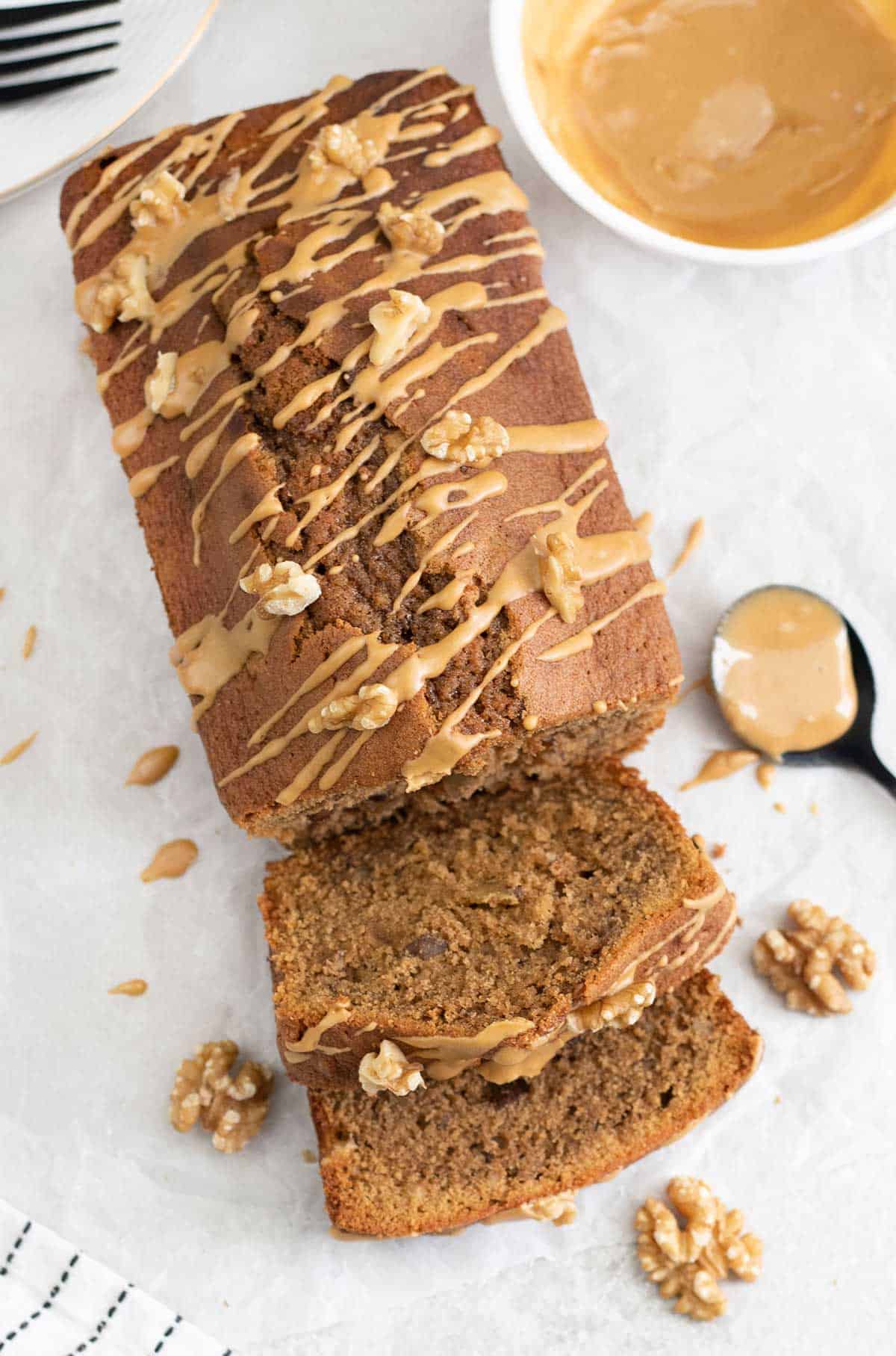 A loaf of banana bread, one of the tastiest loaf pan recipes, topped with a light drizzle of peanut butter and walnut pieces. Two slices are cut from the loaf, with extra walnuts and a spoonful of peanut butter nearby.