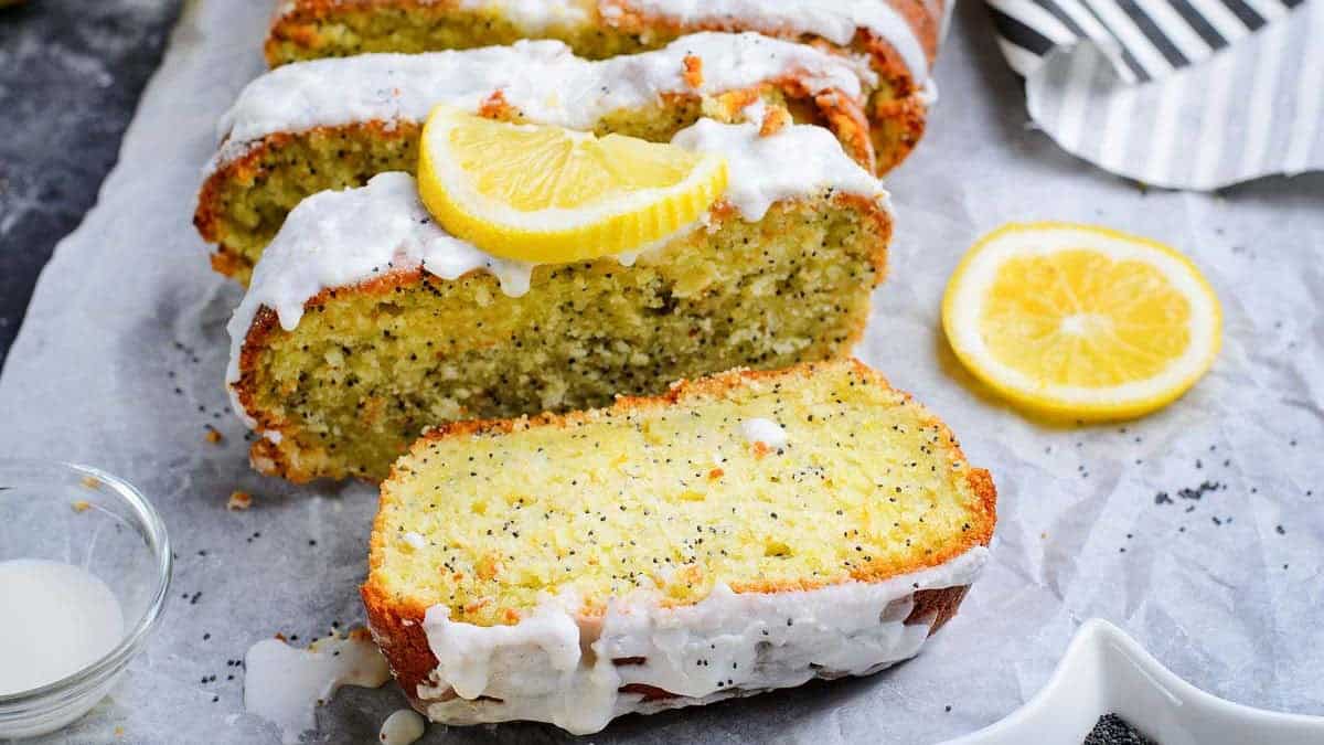 A loaf of lemon poppy seed bread with white icing is sliced, with one slice in the foreground. A lemon wedge and a lemon slice sit on top and beside the loaf, showcasing one of the most delightful loaf pan recipes. Poppy seeds are visible throughout.