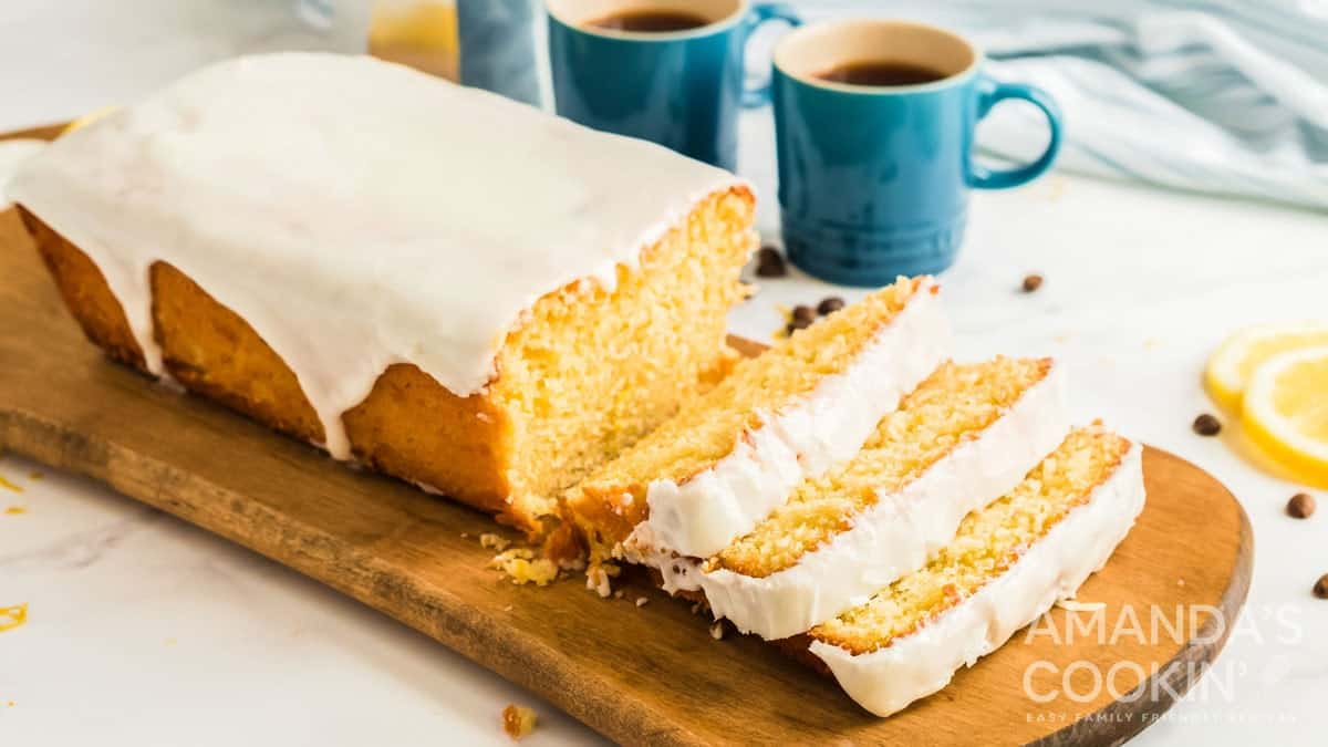 A lemon loaf cake, made with one of our favorite loaf pan recipes, sits on a wooden board. Several slices are cut, revealing a moist, yellow interior. Two blue mugs of coffee and lemon slices are in the background.