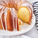 A glazed 7 Up Cake Recipe bundt cake with a slice removed sits on a white plate. The golden brown cake is drizzled with white icing, and a black-and-white striped cloth rests beside it on a white surface.