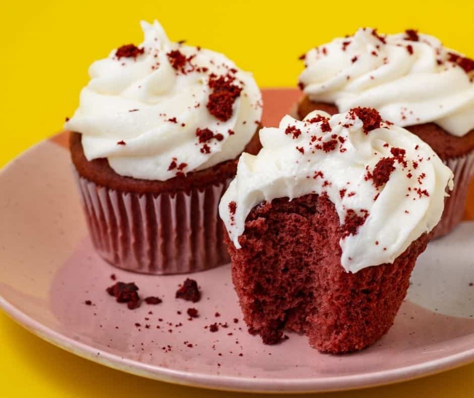 Three red velvet cupcakes with cream cheese frosting, made using Air Fryer Cupcake Recipes, sit on a pink plate against a yellow background; one cupcake has a bite taken out, revealing its moist red interior and crumbs scattered on the plate.