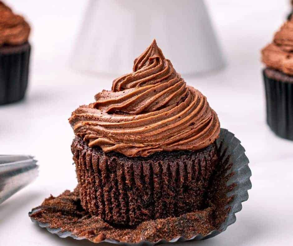 A rich chocolate cupcake with a swirl of chocolate frosting sits in a partially unwrapped black paper liner, a tempting treat inspired by Air Fryer Cupcake Recipes, with more cupcakes and a piping tip visible in the blurred background.