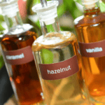 Three glass bottles labeled caramel, hazelnut, and vanilla filled with iced coffee syrups are displayed close together, with green plants blurred in the background.