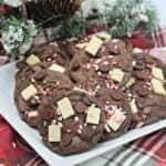 A square white plate holds chocolate cookies made from recipes for cake mix cookies, topped with chocolate chips, white chocolate pieces, and crushed peppermint, set on a red plaid cloth with pine cones and snowy greenery in the background.