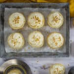 Eight cupcakes with swirled white frosting and gold sprinkles, made from a delightful champagne cupcake recipe, are arranged in a baking tray with parchment paper. Additional cupcakes and a yellow cloth sit nearby on the white marble surface.