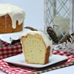 Slice of glazed eggnog cake on a white plate, with the rest of the cake in the background.