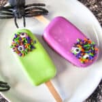 Two popsicles, one green and one purple, decorated with colorful sprinkles and candy eyes, sit on a white plate. Surrounded by plastic black spiders and netting, this fun Halloween setup is perfect for trying creative Cakesicle Recipes.
