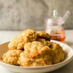 A white plate stacked with golden, fluffy honey butter biscuits sits on a marble surface. In the blurred background, there is a glass jar of honey with a honey dipper inside.