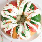 A festive Christmas bundt cake with red, green, and yellow sections, topped with white icing and red, green, and white round sprinkles, sits on a white plate with scattered sprinkles around.