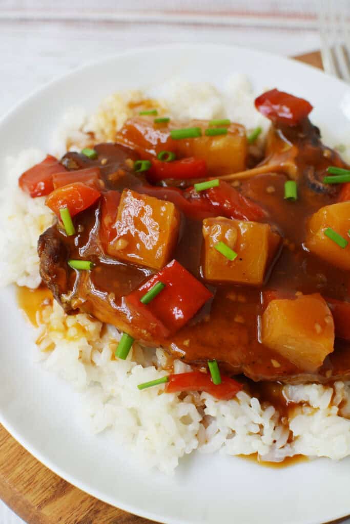 A plate of white rice topped with Hawaiian Pork Chops Slow Cooker style—juicy pork chop, red bell peppers, pineapple chunks, and brown sauce, all garnished with chopped green onions.