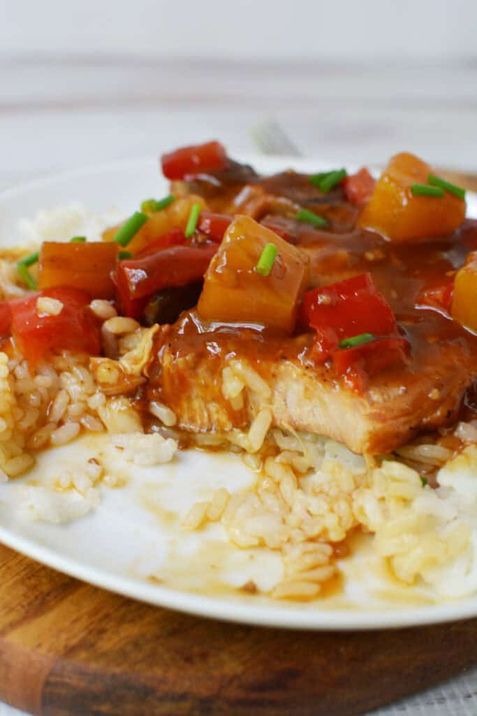 A plate of white rice topped with a piece of salmon covered in a glossy brown sauce, garnished with pineapple, red bell peppers, and green onions—flavors inspired by classic Hawaiian Pork Chops Slow Cooker recipes.