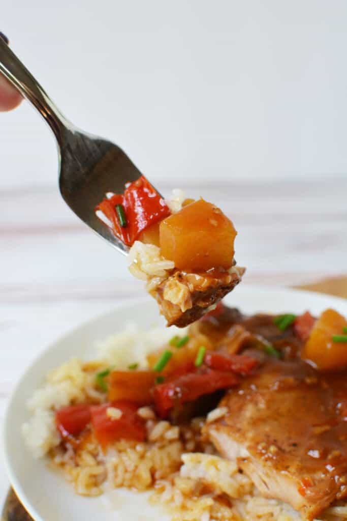 A fork holds a bite of food with rice, red bell pepper, pineapple, and a piece of meat above a plate of similar food, garnished with chopped chives—perfect for serving Hawaiian Pork Chops Slow Cooker style.