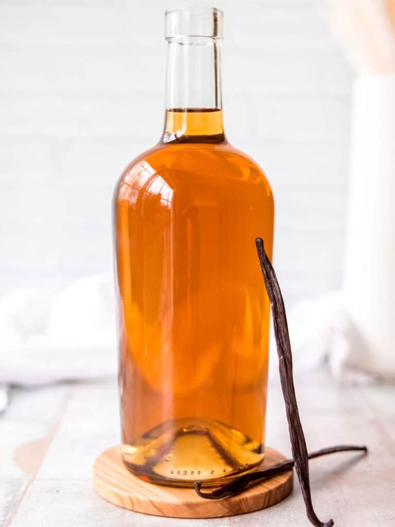 A clear glass bottle filled with amber-colored iced coffee syrup sits on a wooden coaster, with a whole vanilla bean pod leaning against it. The background is softly blurred.