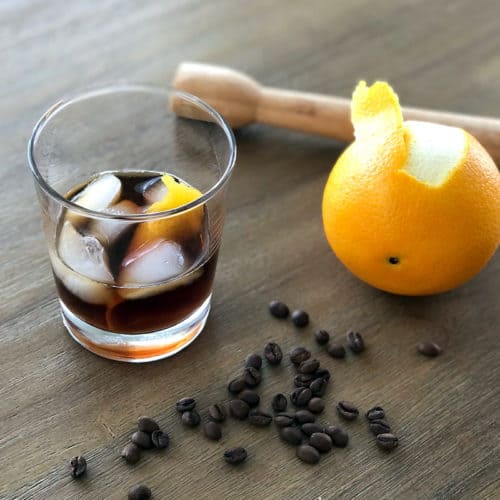 A glass of iced coffee with an orange peel garnish sits on a wooden table next to an orange, a wooden muddler, and scattered coffee beans—inviting you to wonder what alcohol goes well with iced coffee for the perfect pairing.