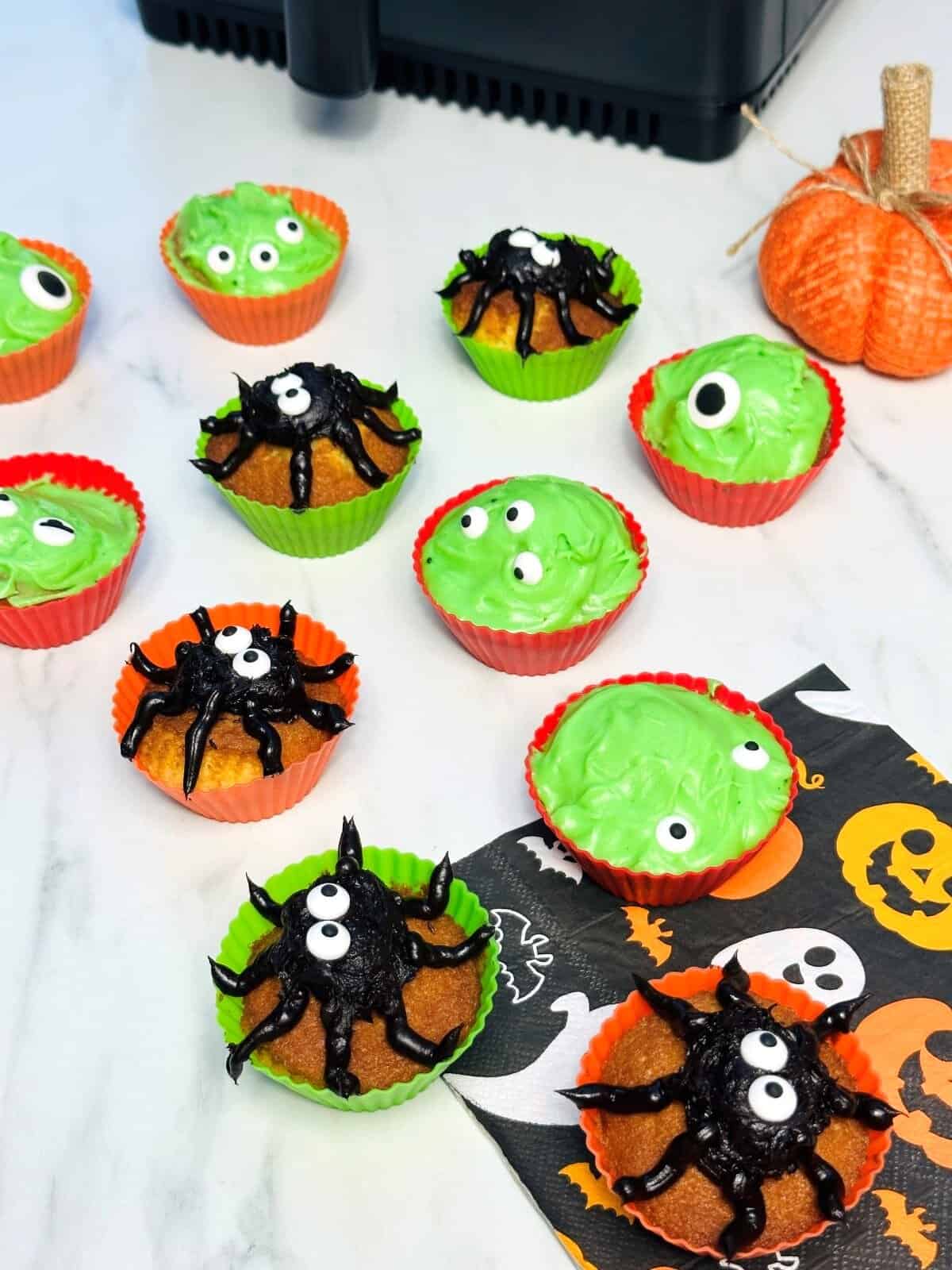 Halloween-themed cupcakes in colorful liners, inspired by Air Fryer Cupcake Recipes, are topped with green frosting and candy eyes or black icing spiders. A small decorative pumpkin and a festive napkin sit nearby on a white surface.