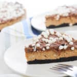 A slice of chocolate cheesecake with a cookie crust and mini marshmallows on top sits on a white plate, making it the perfect no bake Christmas treat, with a fork in front and more cheesecake slices in the background.