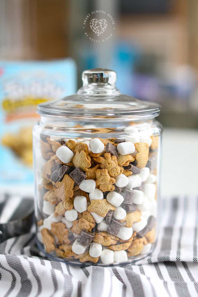 A glass jar filled with mini marshmallows, chocolate chunks, and small bear-shaped graham crackers—perfect for campfire snacks—sits on a striped cloth in a bright kitchen setting. The jar has a lid, ready to keep treats fresh.