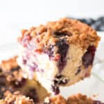 A close-up of a slice of blueberry coffee cake with a crumbly streusel topping, being lifted from a baking dish. Perfect for fans of 9x13 cake recipes, the cake appears moist with visible blueberries throughout.