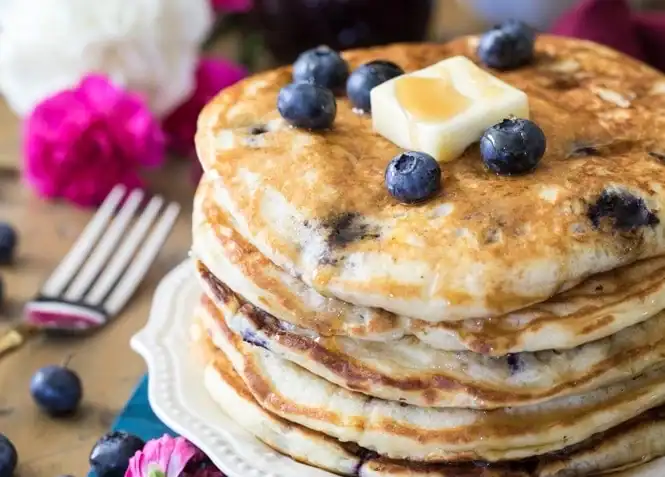 A stack of blueberry pancakes topped with fresh blueberries, a pat of butter, and syrup on a white plate—perfect for those searching for delicious frozen blueberry recipes. A fork and pink flowers sit nearby.