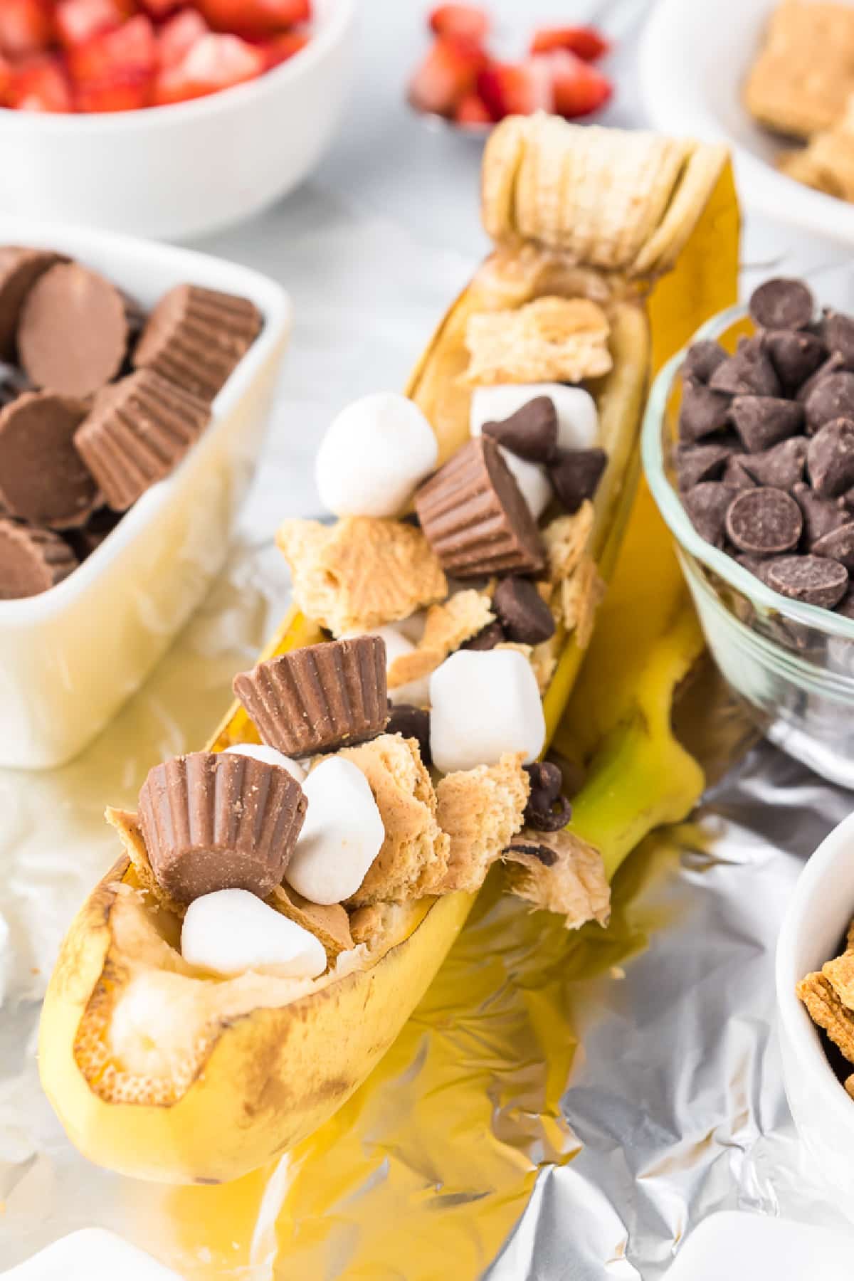 A banana split open and filled with mini marshmallows, chocolate chips, graham cracker pieces, and mini peanut butter cups—this campfire snack is surrounded by bowls of these ingredients on a sheet of foil.