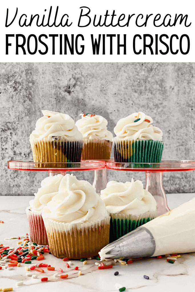 Cupcakes with vanilla Crisco Frosting Recipe buttercream and colorful sprinkles are displayed on a pink cake stand, with a piping bag and more sprinkles scattered on a white surface in front. Text above reads Vanilla Buttercream Frosting With Crisco.