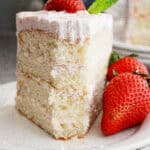 A slice of layered vanilla cake with white frosting sits on a white plate, garnished with two fresh strawberries and a mint leaf. The partially eaten cake has a light, fluffy texture—perfect for trying out a strawberry champagne cake recipe.