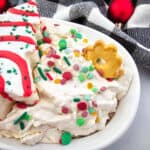 A white bowl of festive holiday dip, one of the easiest No Bake Christmas Treats, is topped with colorful sprinkles and served with a red-and-green iced Christmas tree cookie and a small wreath cookie, set against a checkered cloth and red ornaments.