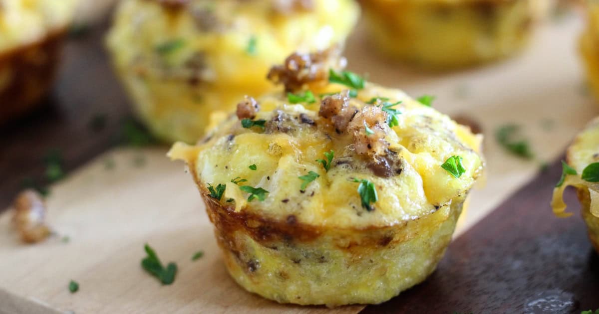 Mini egg muffins topped with melted cheese, sausage crumbles, and chopped parsley, displayed on a wooden surface.