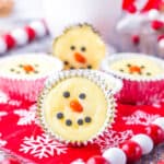 Mini cheesecakes decorated as snowman faces with chocolate chips and candy for features, displayed on a red snowflake-patterned napkin—these are the perfect cute Christmas desserts, accented by festive red and white beads in the background.