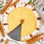 A festive No Bake Eggnog Pie with a slice missing sits on a table, topped with whipped cream and star anise, surrounded by cinnamon sticks, greenery, and holiday-themed napkins.