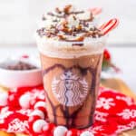 A festive Starbucks chocolate frappuccino, inspired by Starbucks drink recipes, is topped with whipped cream, chocolate drizzle, and a candy cane. It sits on a wooden board with a red snowflake napkin and holiday decorations in the background.