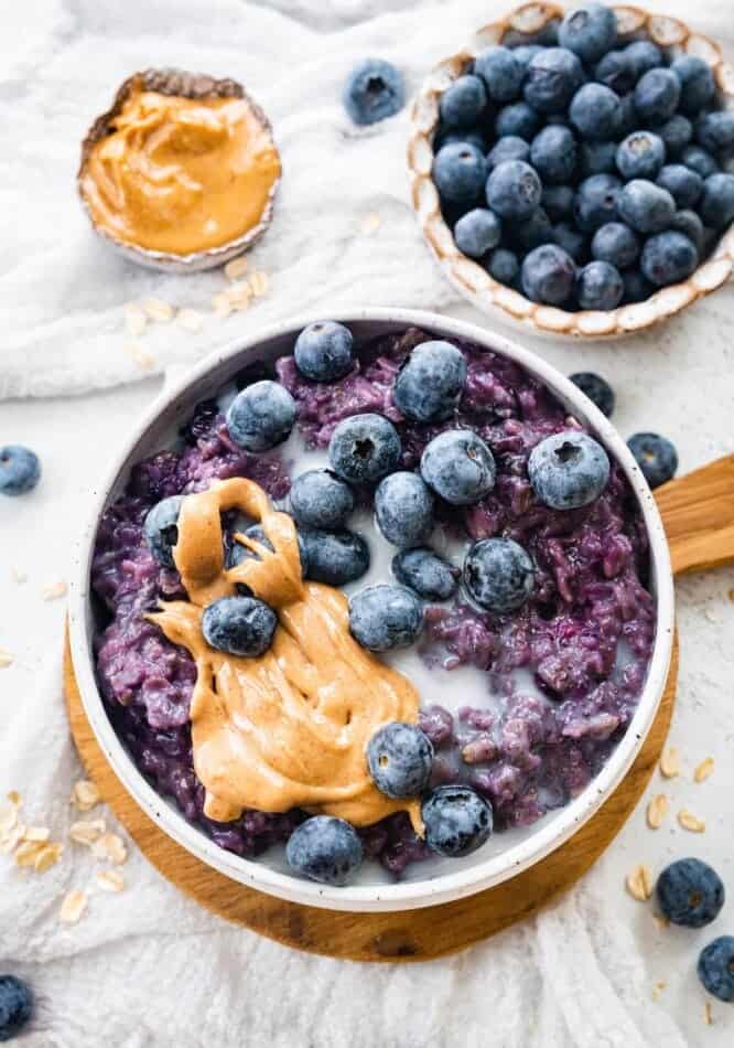 A bowl of blueberry oatmeal topped with fresh blueberries, a swirl of peanut butter, and a splash of milk sits on a wooden board—perfect inspiration for frozen blueberry recipes. Nearby are scattered oats, more blueberries, and a halved coconut shell.