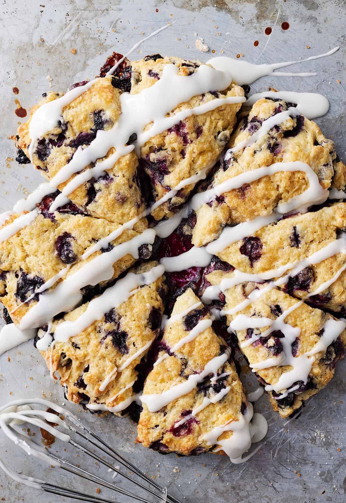 Triangular blueberry scones—a delicious twist on frozen blueberry recipes—are topped with white icing drizzle and arranged closely together on a metal baking sheet, with a whisk and icing traces visible nearby.