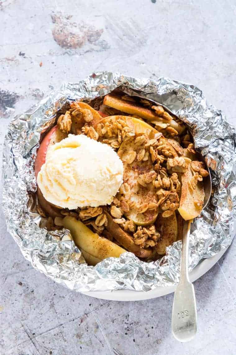 A bowl lined with foil holds baked apple slices topped with granola and a scoop of vanilla ice cream, making for the perfect campfire snack, with a spoon resting on the side.