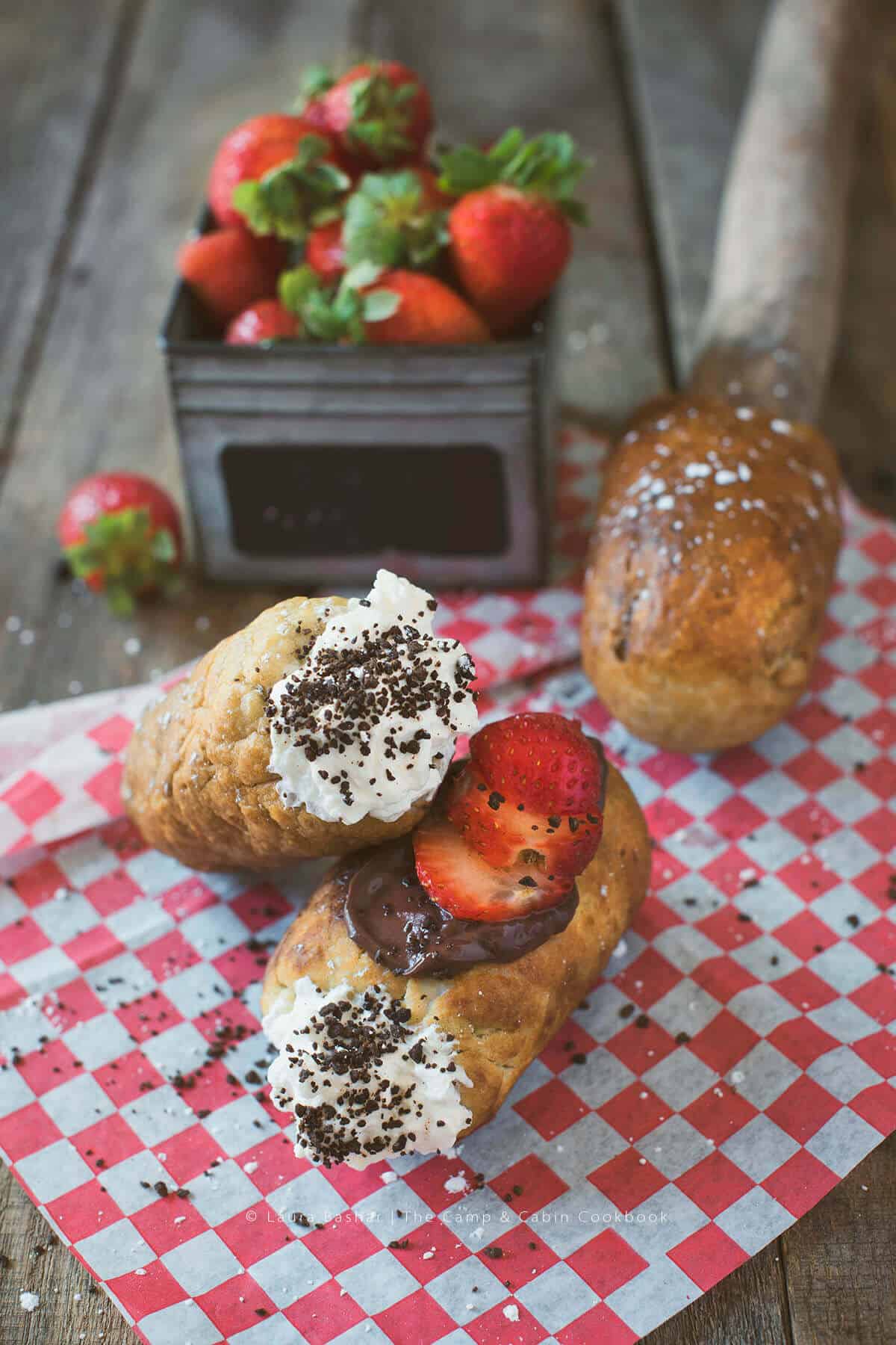 Two stuffed eclairs, perfect for campfire snacks, are topped with whipped cream, chocolate, sprinkles, and fresh strawberry slices on red-checkered paper, with a box of strawberries and a loaf of bread in the background.