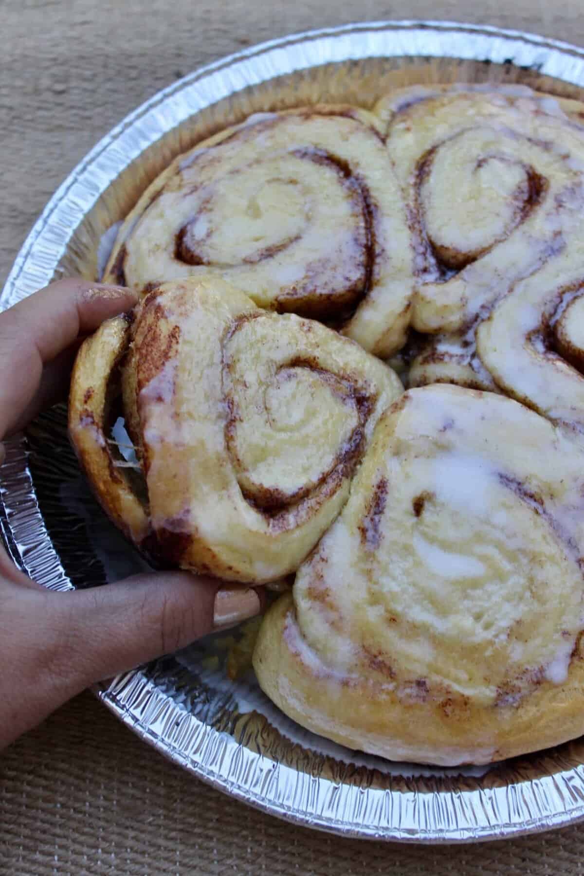 A hand lifts a cinnamon roll from a round foil tray filled with iced cinnamon rolls, set on a textured brown surface—perfect campfire snacks for sharing under the stars.