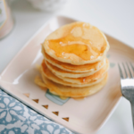 A small stack of pancakes drizzled with syrup on a square plate beside a fork.