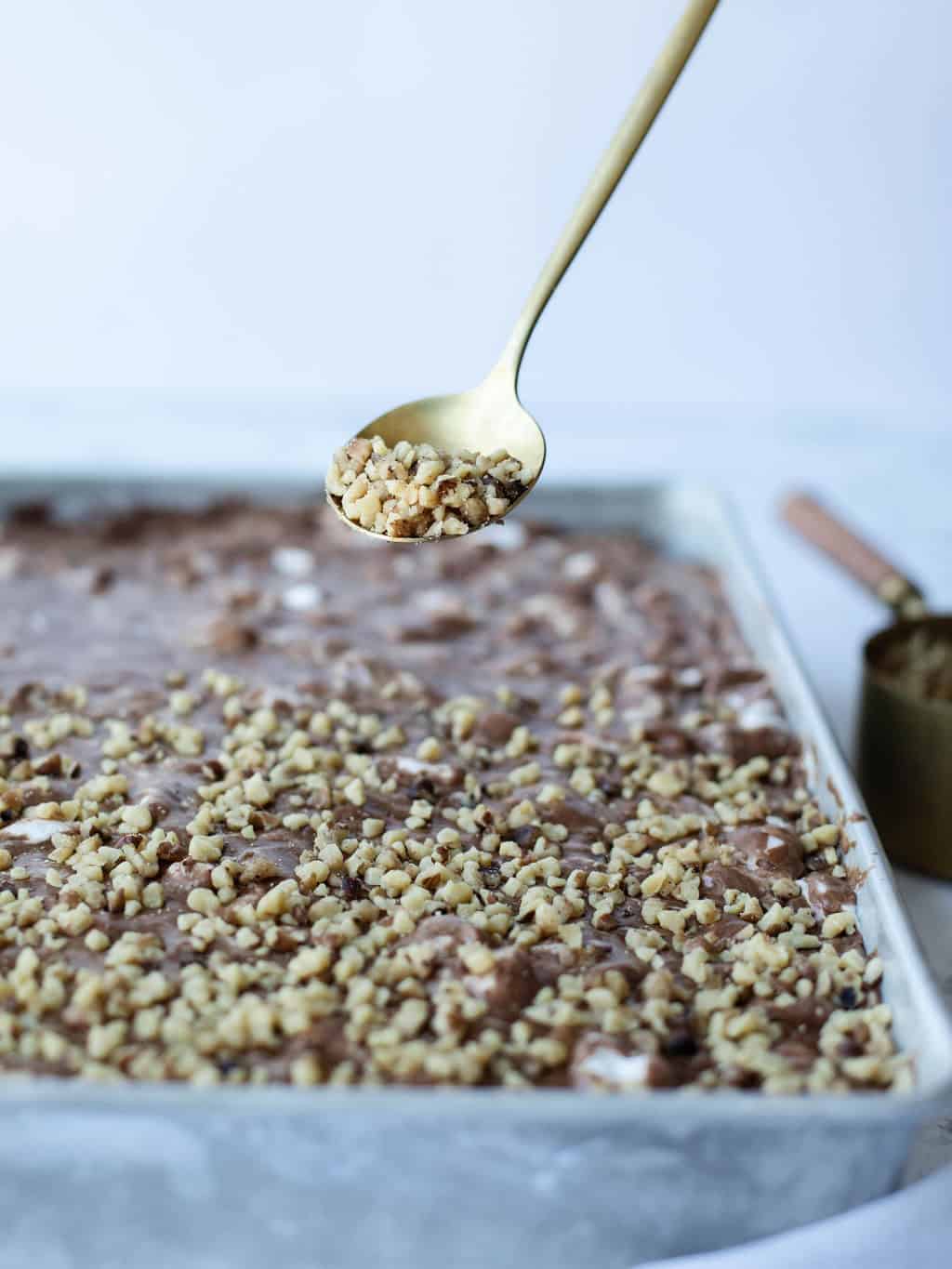 A close-up of a spoon sprinkling chopped nuts over a tray of chocolate dessert batter, perfect for 9x13 cake recipes, with more nuts visible in a measuring cup in the background.
